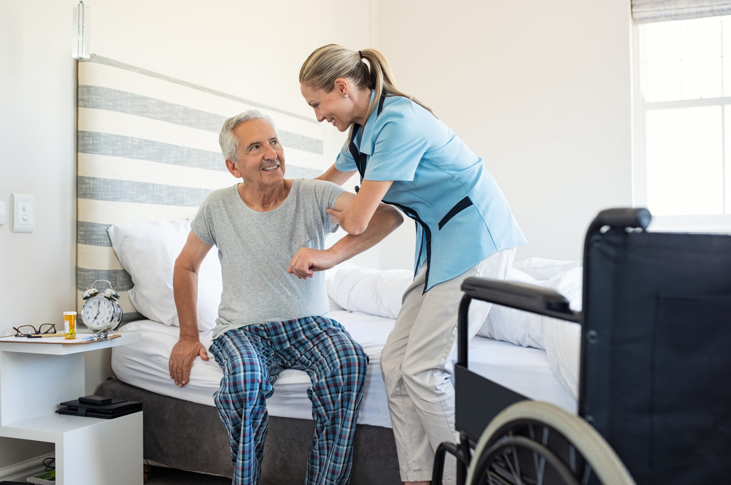 Smiling nurse assisting senior man to get up from bed. Caring nurse supporting patient while getting up from bed and move towards wheelchair at home. Helping elderly disabled man standing up in his bedroom.